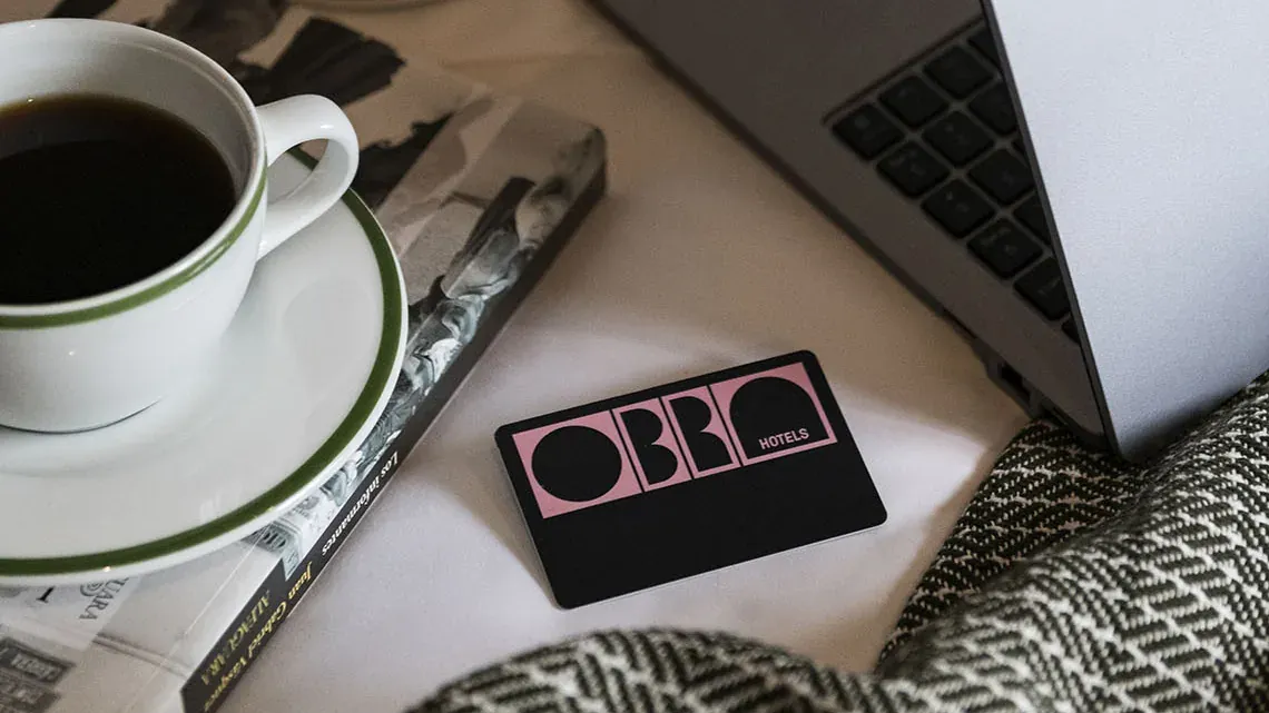Café, revista, portátil y tarjeta llave rosada de OBRA Hotels sobre la cama.